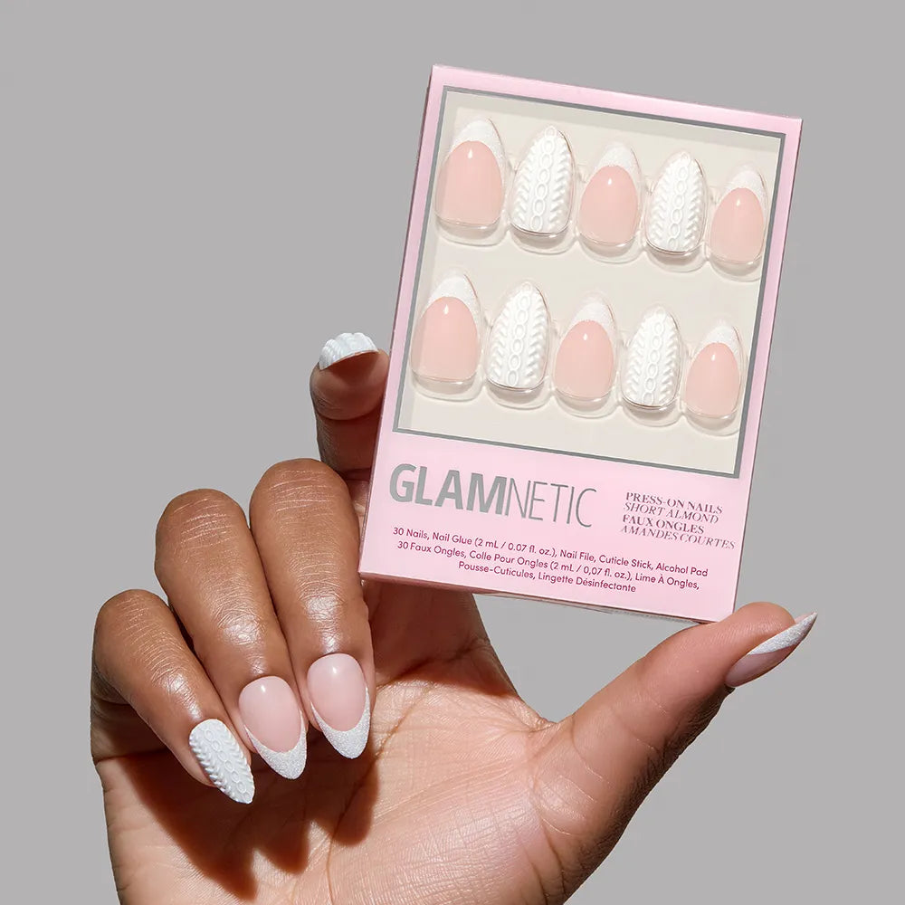 A hand with manicured short almond nails featuring a pink base and white textured designs holds a box of glamnetic Sweater Weather, displaying matching pink and white patterned nails against a plain gray background.