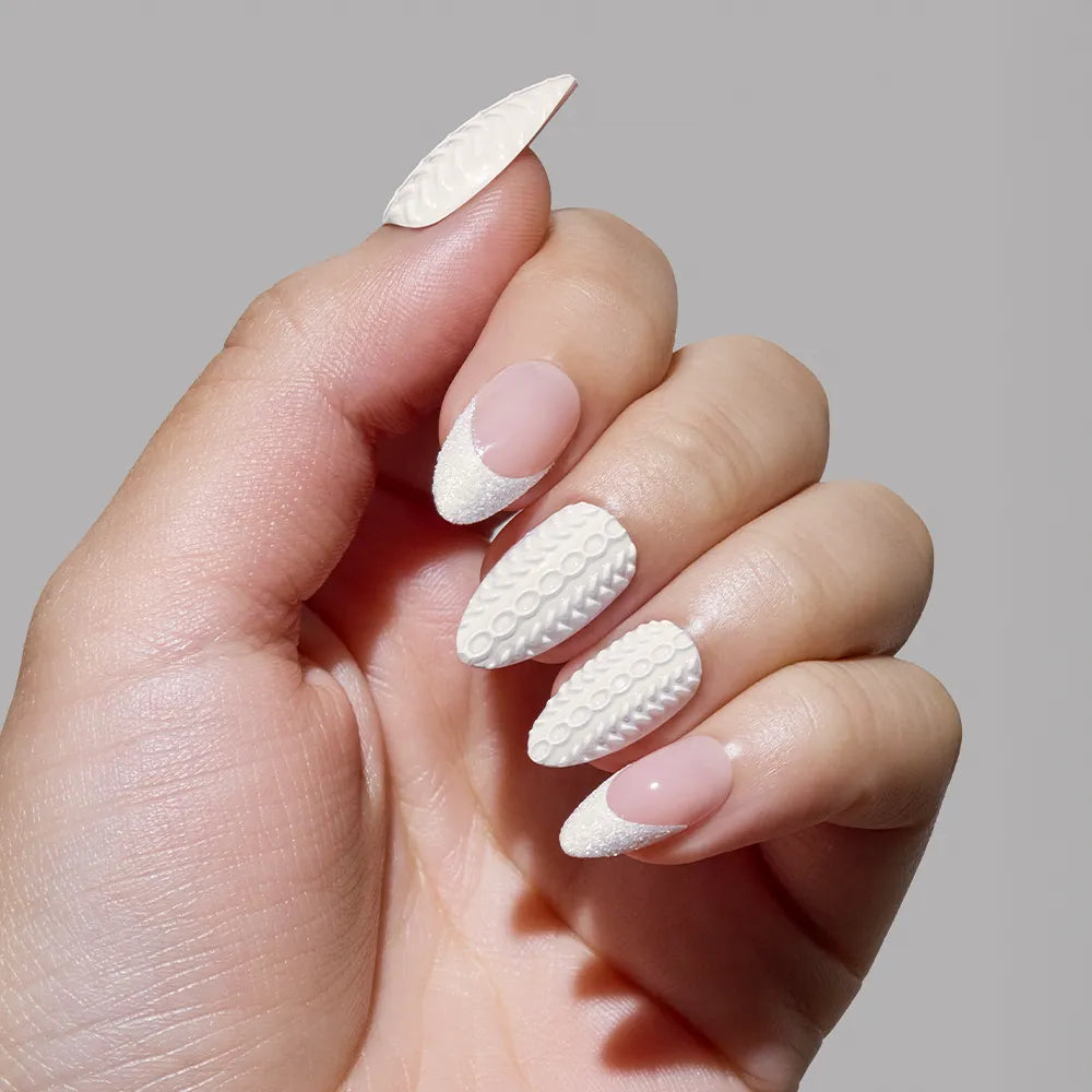 A hand with short almond nails in a nude base and white textured sweater-inspired designs showcases the glamnetic Sweater Weather set against a plain gray background. - Product image
