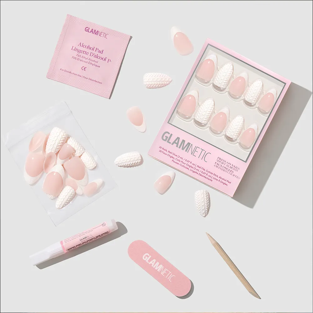 A flat lay of the glamnetic Sweater Weather Nail Set shows short almond nails with pink and white textured designs, glitter French tips, glue, a pink nail file, wooden stick, alcohol pad, and a resealable bag on a light surface. - Product image