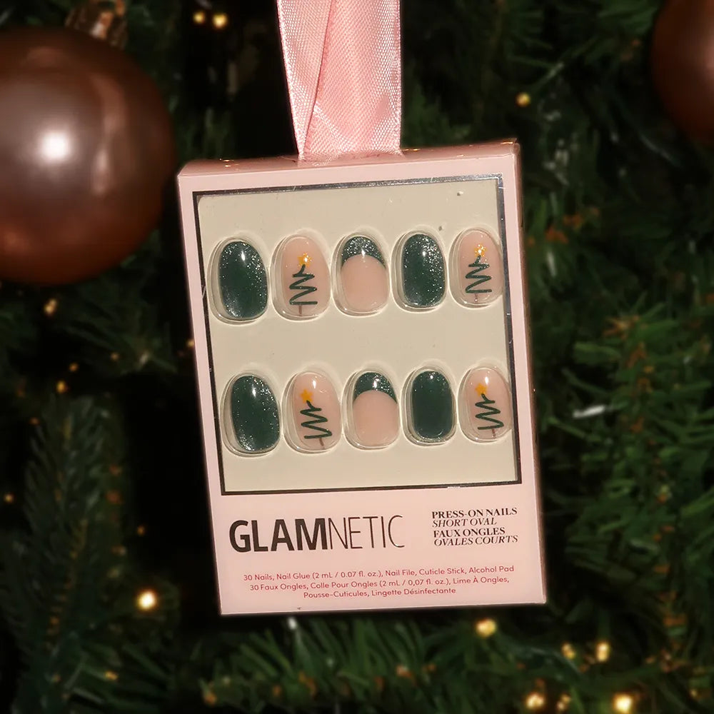 A box of glamnetic Velvet Pine nails hangs on a pink ribbon before a festive Christmas tree. This holiday set features green nails, some adorned with simple Christmas tree and candle designs. - Product image