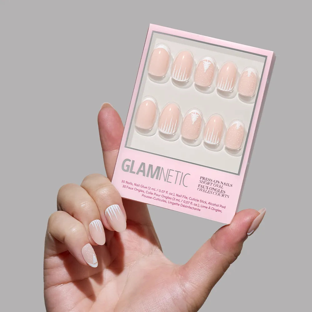 A hand with light pink and white striped French tip press-on nails holds a box of glamnetic Winter's Tale press-on nails featuring a pink and white geometric design, perfect for winter nail art, against a plain gray background.