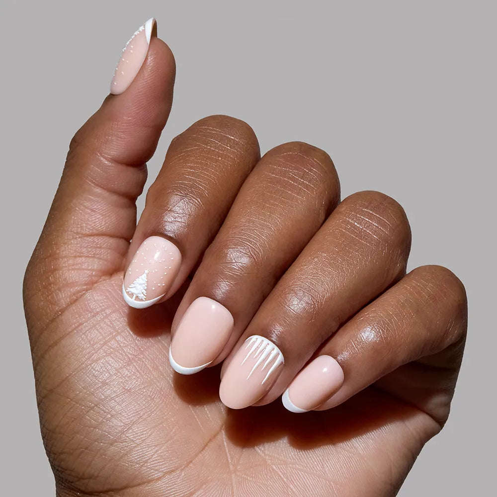 A hand with medium brown skin displays glamnetic's Winter's Tale press-on nails: short, almond-shaped and nude, featuring minimalist white winter nail art like dots, lines, a tree, and French tips, against a plain gray background. - Product image