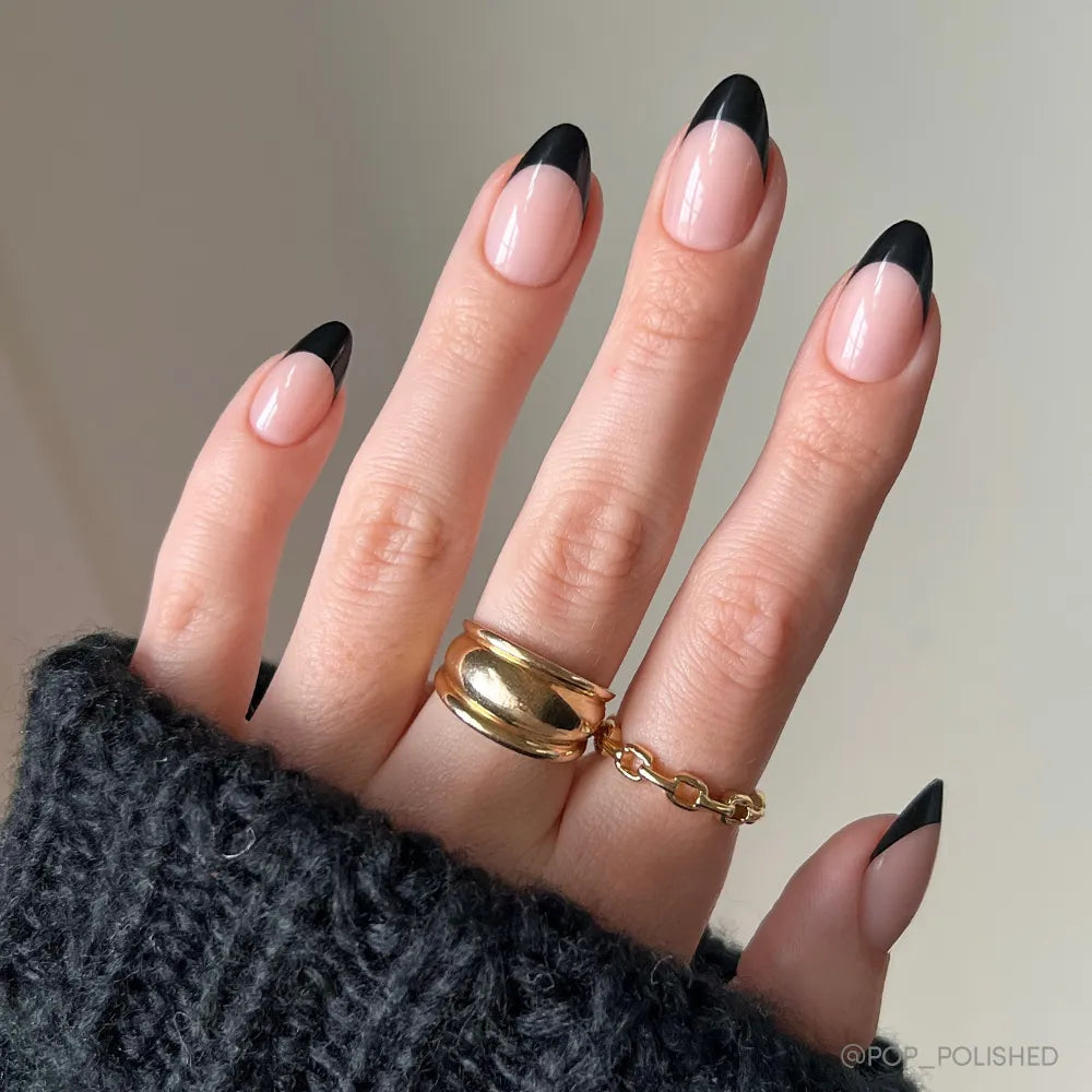 A hand with short almond nails in glamnetic's Caviar press-ons, styled with modern black French tips. Chunky gold rings and a black knit sweater highlight the chic glamour of these elegant nails.