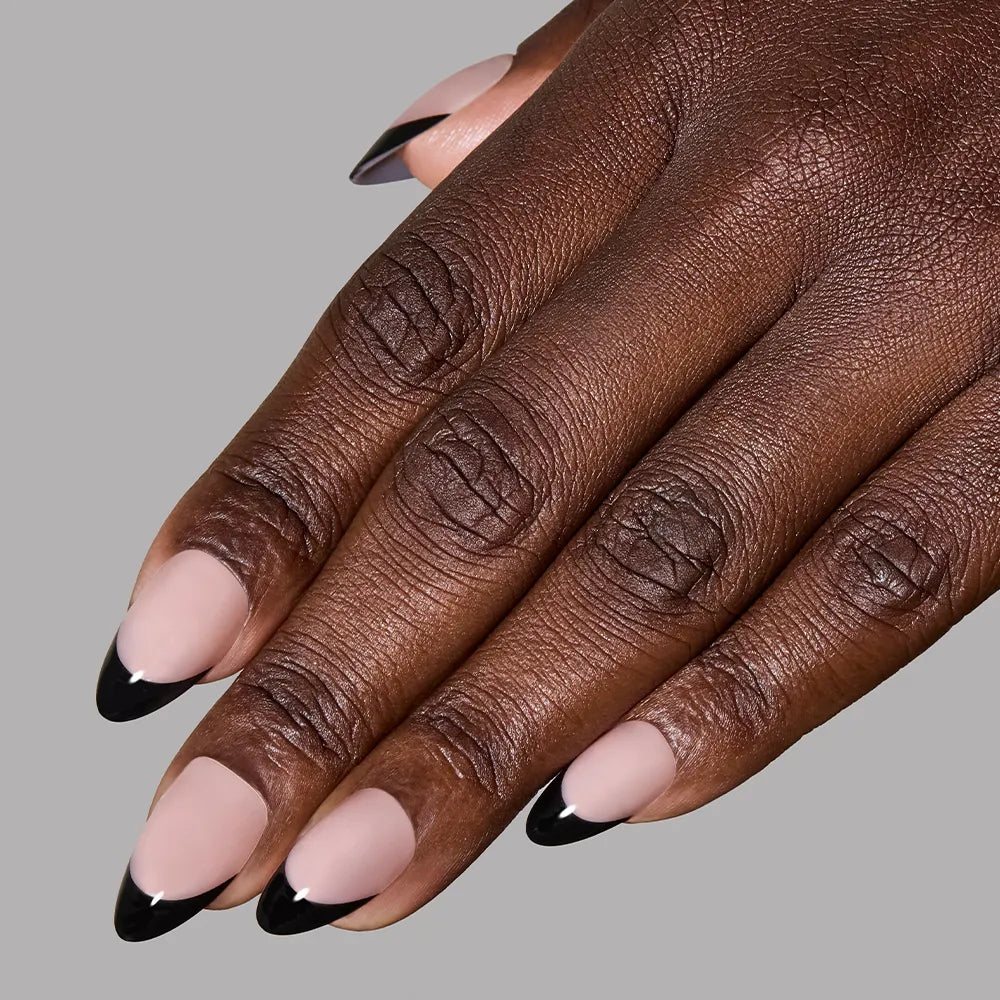A close-up of a hand with short almond nails showcases glamnetic's Caviar press-ons, featuring a nude base and glossy black French tips, set against a plain gray background. - Product image