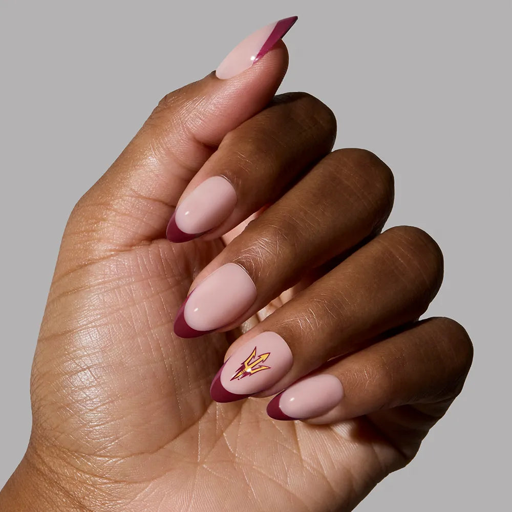 A hand with almond-shaped glamnetic Arizona State Sun Devils Press-On Nails featuring a nude pink base, maroon French tips, and a gold lightning bolt on the ring finger, shown against a plain gray background. - Product image