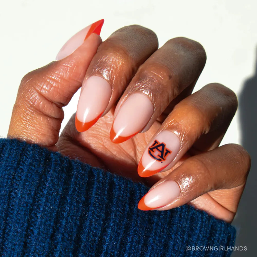 A hand wears glamnetic's Auburn Tigers Press-On Nail Set, with almond-shaped nude nails and bright orange French tips. The ring finger features the team's blue and orange logo. A blue knit sleeve is worn. - Product image