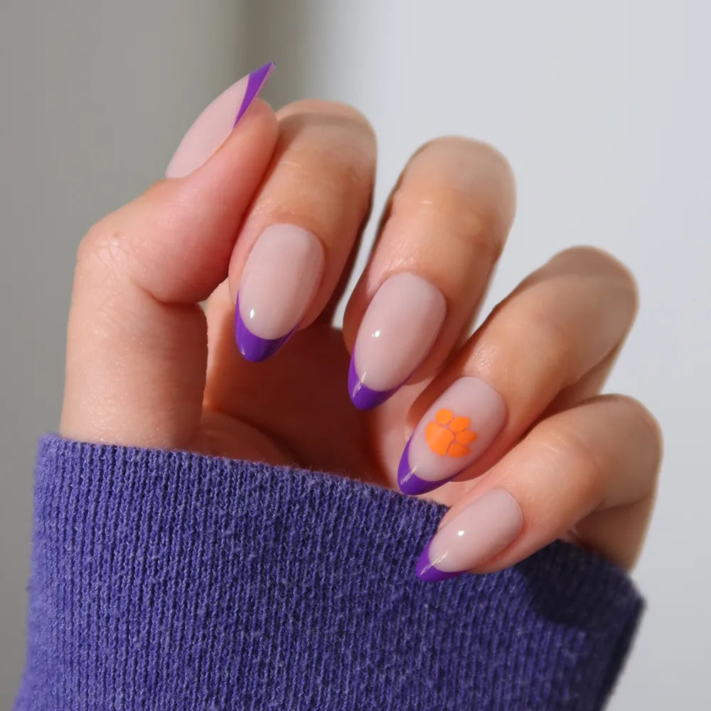 The glamnetic Clemson Tigers Press-On Nail Set features long almond-shaped nude nails with purple French tips and an accent nail with an orange flower, perfect for showing team spirit. One nail is pictured with a purple sleeve. - Product image