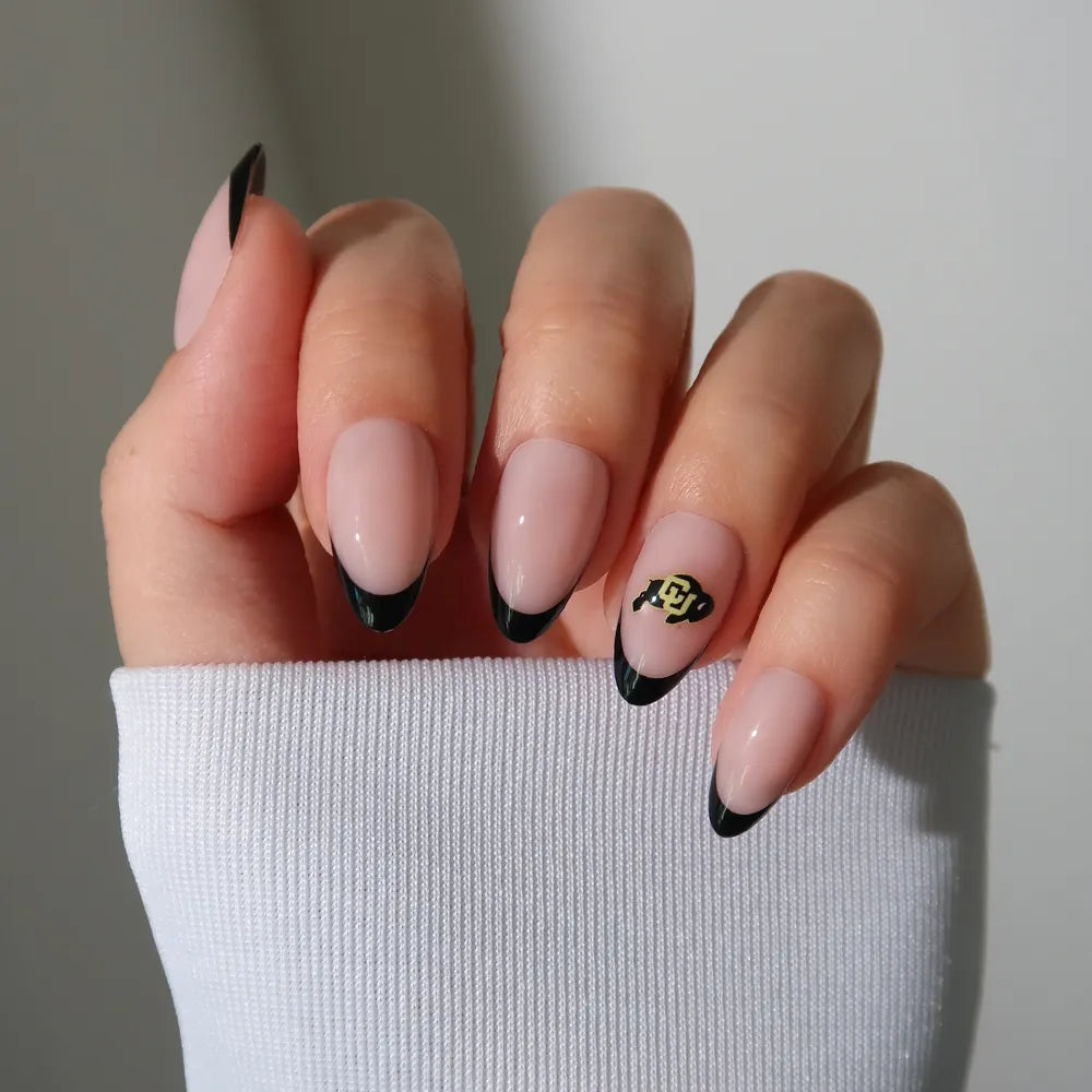 A hand with glamnetic’s Colorado Buffaloes Press-On Nail Set—black French tips on a nude base and a gold/black team logo accent—peeks from beneath a white sleeve, highlighting the salon-quality manicure. - Product image