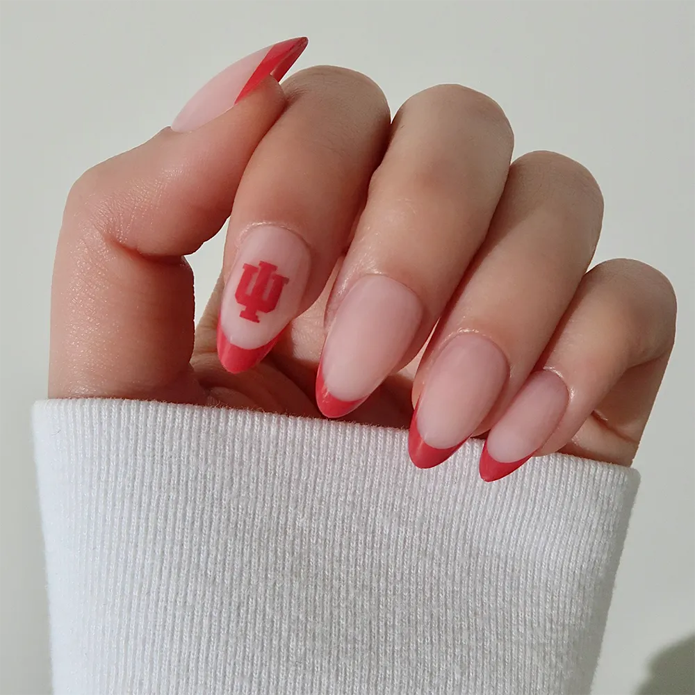 A hand wears the glamnetic Indiana Hoosiers Press-On Nail Set: almond-shaped nails with a pink base, crimson French tips, and an IU logo on the ring finger. The person is dressed in a white ribbed sleeve. - Product image