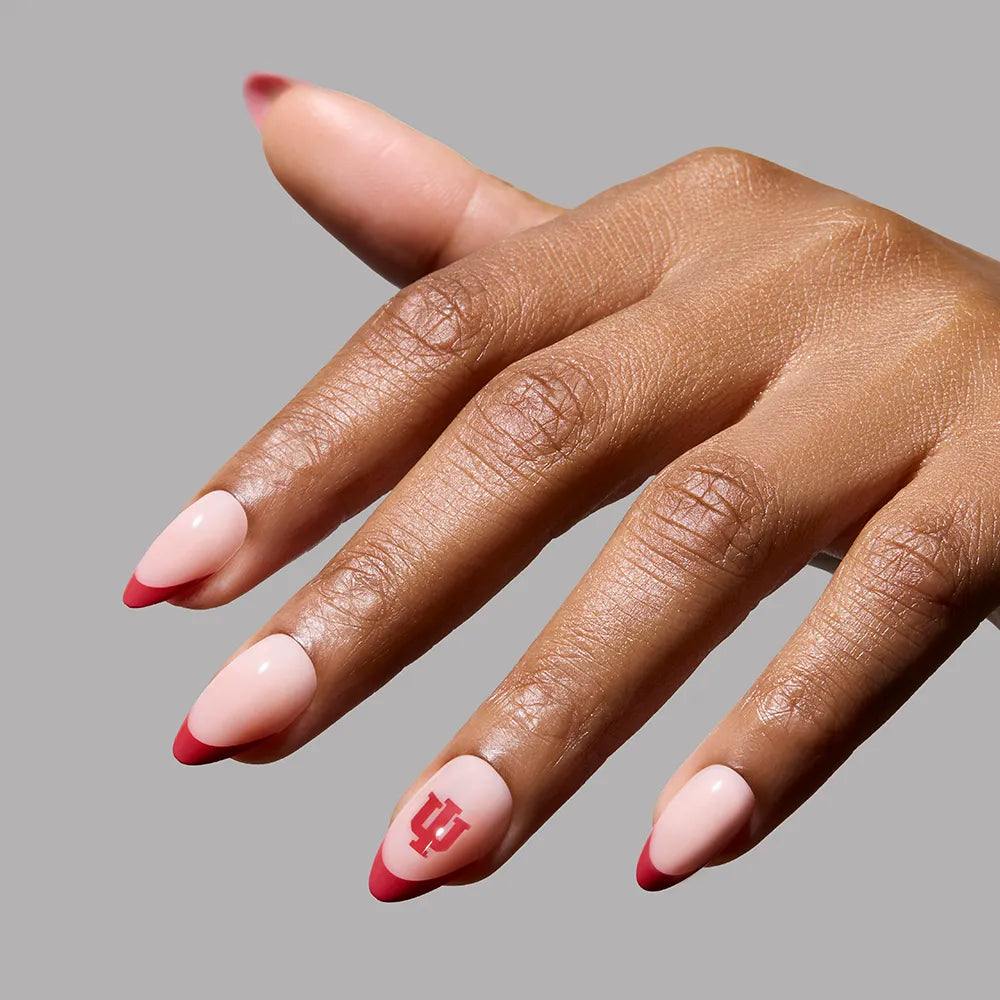A hand with almond-shaped crimson French tip nails in pale pink, featuring the Indiana University logo on the ring fingernail, showcases the glamnetic Indiana Hoosiers Press-On Nail Set against a solid gray background. - Product image