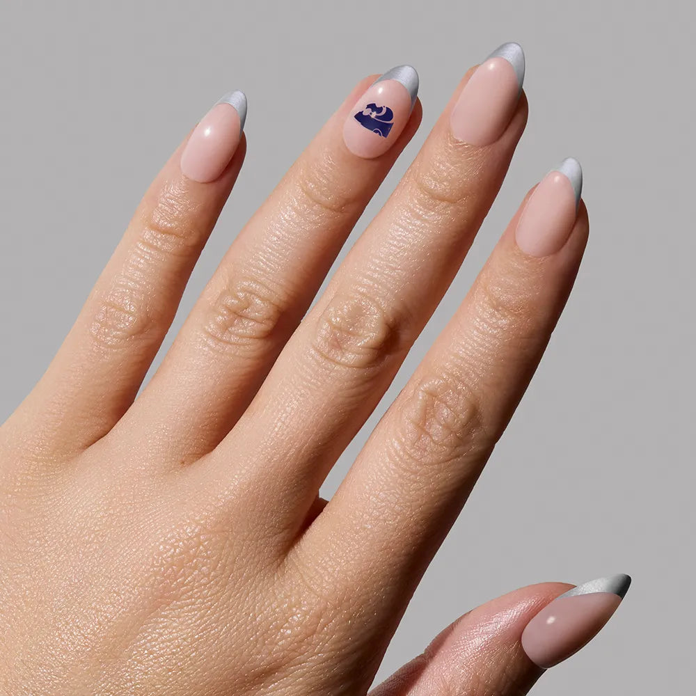 A hand with almond-shaped Kansas State Wildcats Press-On Nails from glamnetic. The nails are painted nude with silver tips, and the ring finger features a dark blue Aquarius zodiac symbol. Background is plain light gray. - Product image