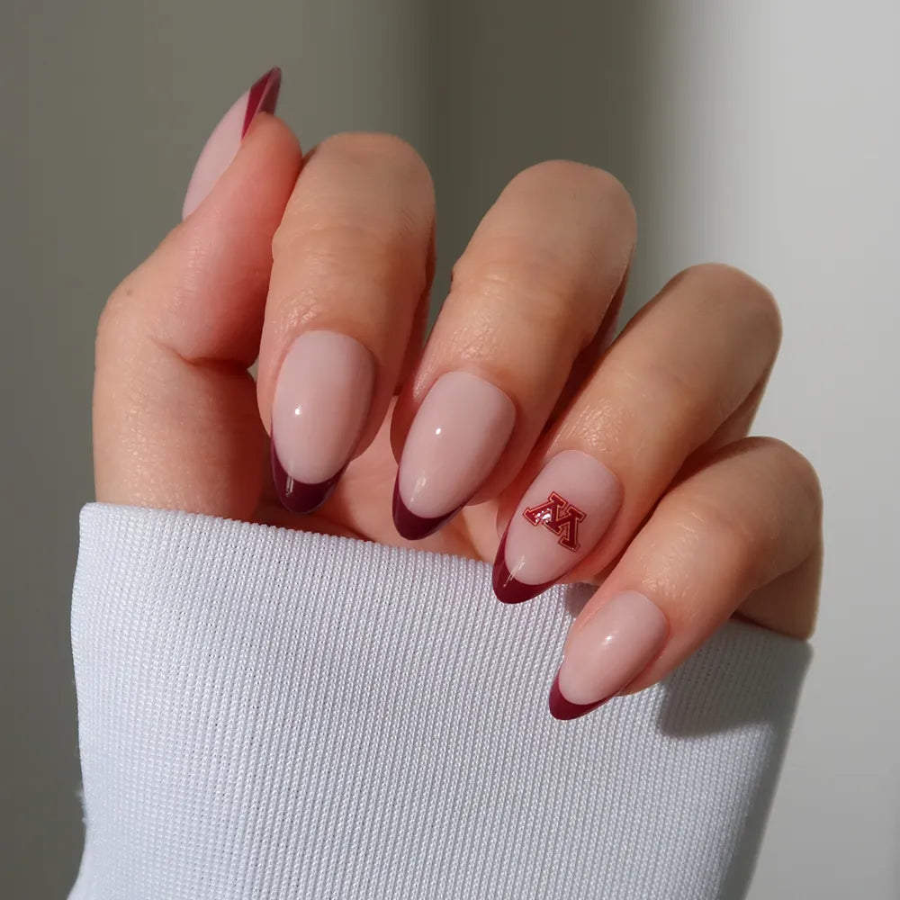 Show off your game-day style with glamnetic’s Minnesota Golden Gophers Press-On Nail Set: almond-shaped nails, nude base, dark red French tips, and a red M sticker on the ring finger. Finished with a white long-sleeve shirt for a classic look. - Product image