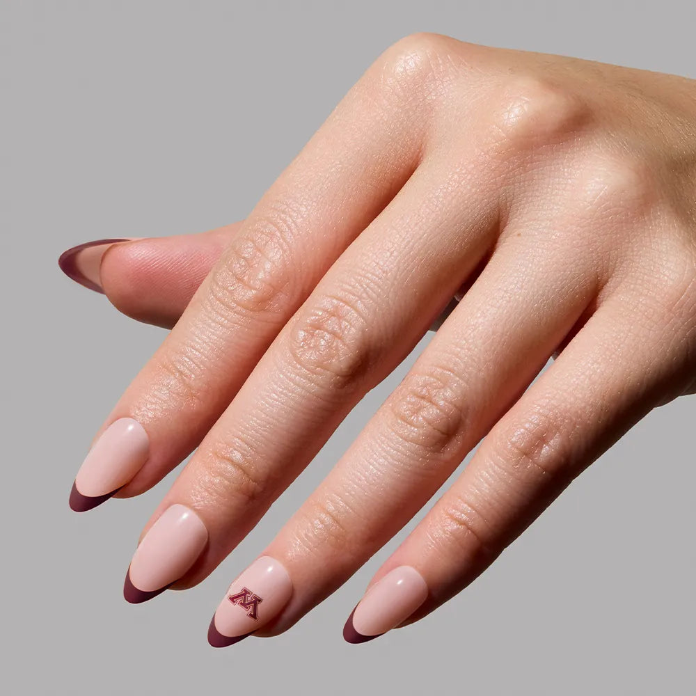 A hand models the glamnetic Minnesota Golden Gophers Press-On Nail Set, featuring almond-shaped nude pink nails with brown tips and a small red geometric design on the ring finger, perfect for a subtle game-day look against a gray background. - Product image
