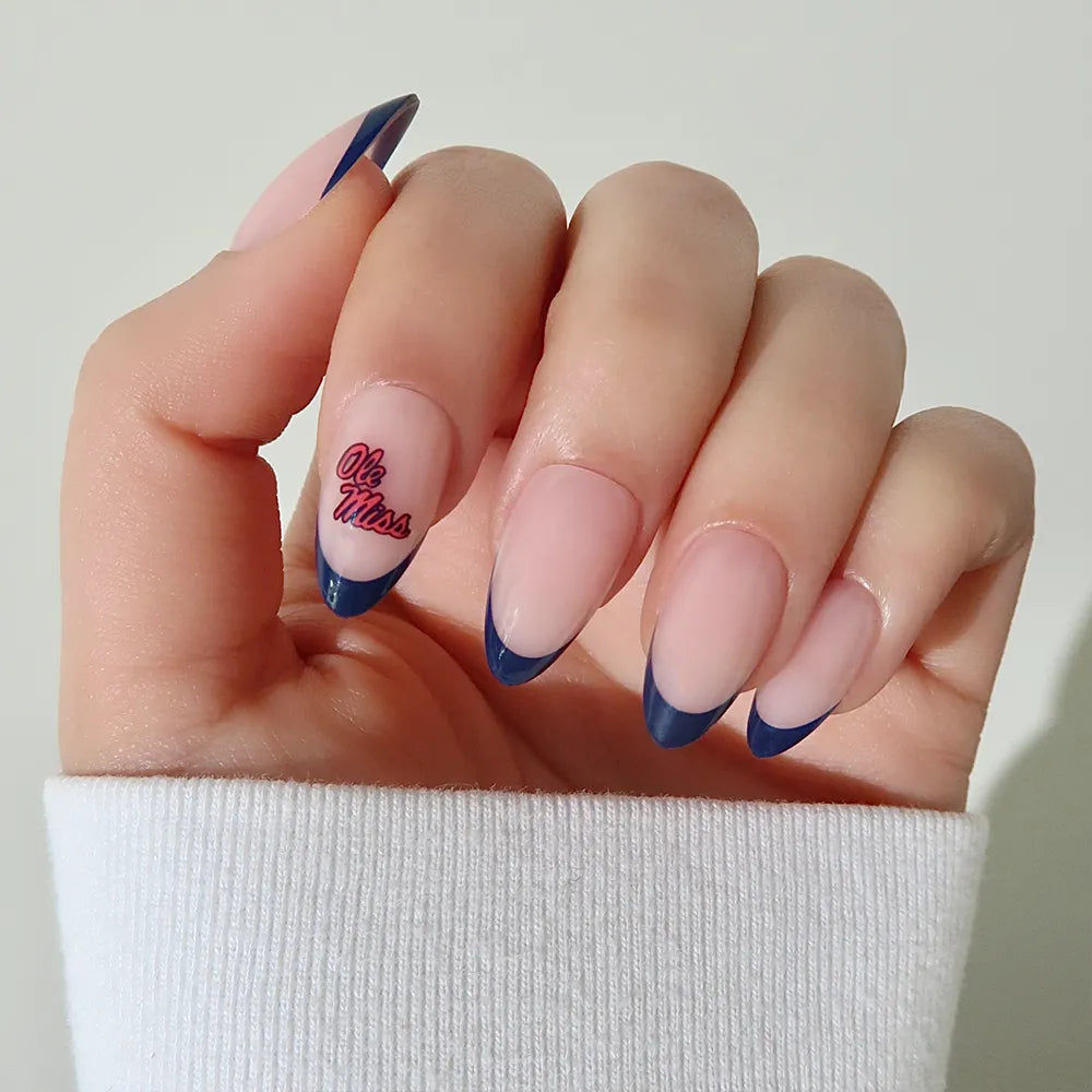 A hand wears the glamnetic Mississippi Rebels Press-On Nail Set, showing almond-shaped, salon-quality nails with a nude base, dark blue French tips, and "Ole Miss" in red and blue script. A white long-sleeve shirt completes the look. - Product image