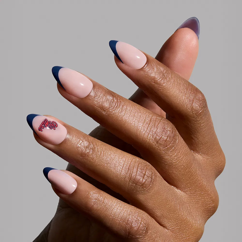 A hand wears glamnetic’s Mississippi Rebels Press-On Nail Set: almond-shaped nails in nude with dark blue French tips and red cursive on one, set against a plain light gray background. - Product image