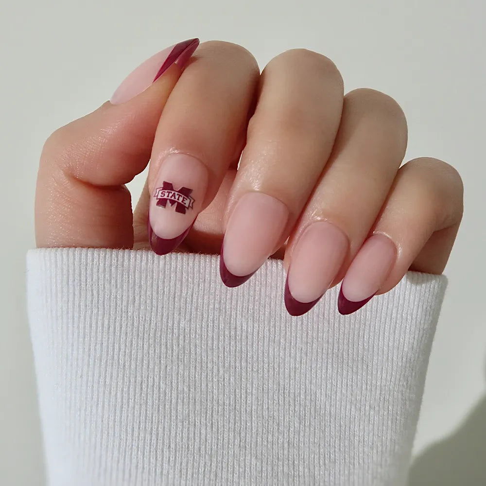 A hand with long almond-shaped nails showcases glamnetic's Mississippi State Bulldogs Press-On Nail Set, featuring matte nude polish, maroon French tips, a Bulldogs decal on the ring finger, and a white sleeve partially covering the hand. - Product image