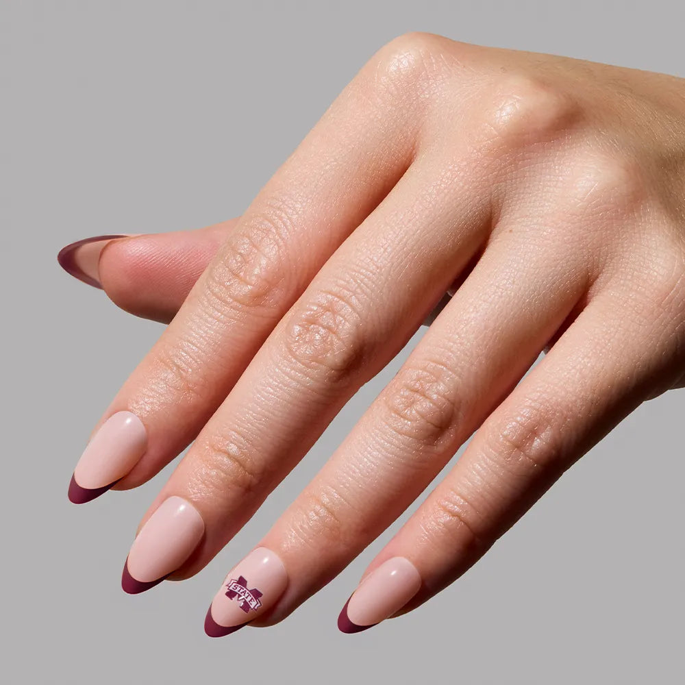 A hand displays the glamnetic Mississippi State Bulldogs Press-On Nail Set—almond-shaped nude nails with bold maroon French tips and a small decorative detail—against a plain light gray background. - Product image