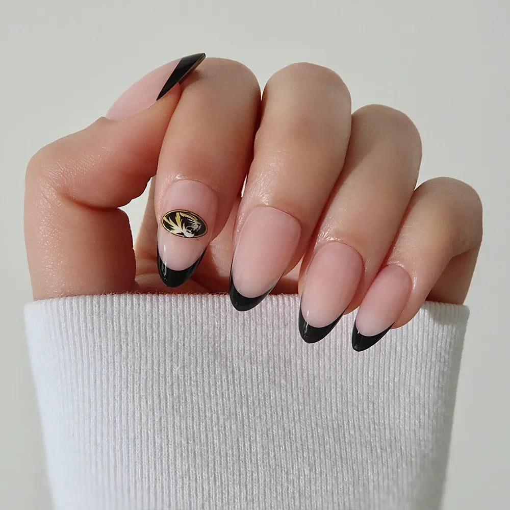 The Missouri Tigers Press-On Nail Set by glamnetic features almond-shaped nails with a nude base, black French tips, and a gold-black leaf accent—ideal for subtle team logo vibes. Shown with a white long-sleeve top. - Product image
