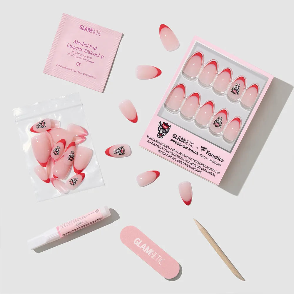 The glamnetic NC State Wolfpack Press-On Nail Set includes pink and red nails, a pink nail file, wooden cuticle stick, alcohol pad, glue tube, and extra nails—all arranged on a light gray surface. - Product image