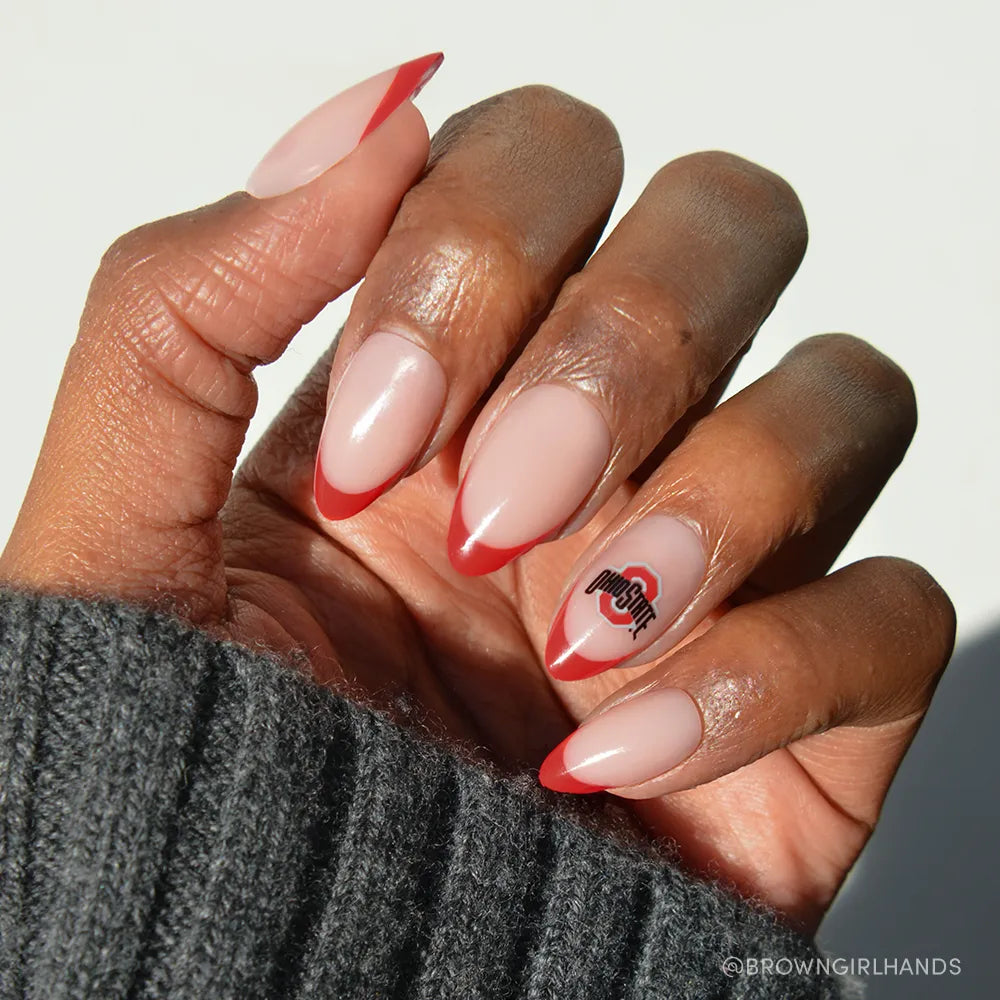 A hand wears the glamnetic Ohio State Buckeyes Press-On Nail Set with almond nude nails, red French tips, and the Ohio State logo on the ring finger—perfect game-day manicure—shown against a gray knit sleeve. - Product image