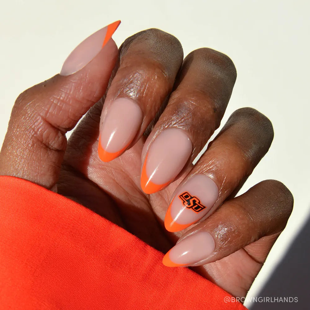 A hand with almond-shaped glamnetic Oklahoma State Cowboys Press-On Nails: nude base, bold orange French tips, and an OSU logo accent nail—perfect for game day. An orange sleeve completes the spirited look. - Product image