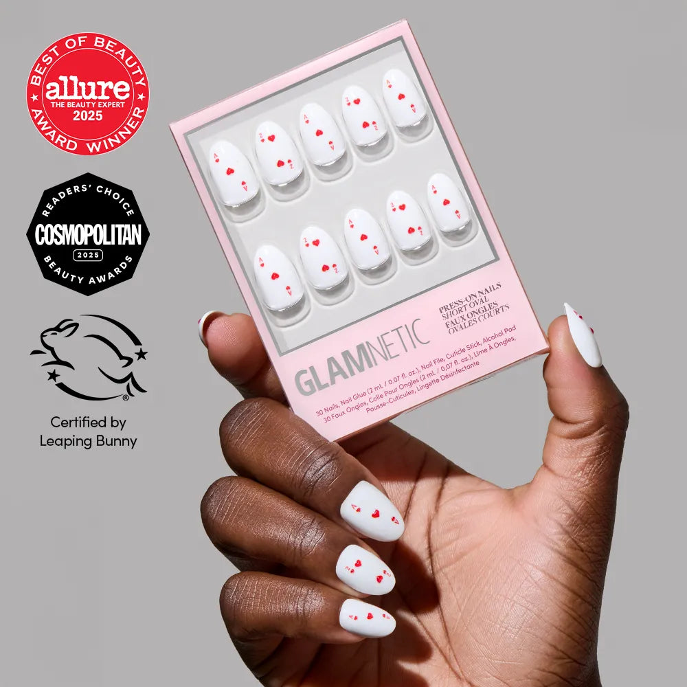 A hand with oval, white press-on nails from glamnetic's Ace of Hearts set—decorated with small red hearts—holds the matching box. Award logos and a Leaping Bunny Certified symbol are shown on the left.