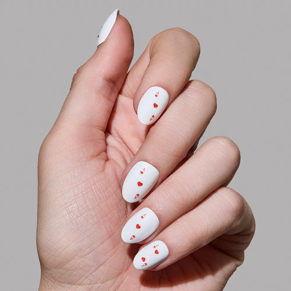 A hand displays glamnetic's Ace of Hearts press-on nails—long, oval, white nails adorned with small red heart designs—against a plain gray background. - Product image