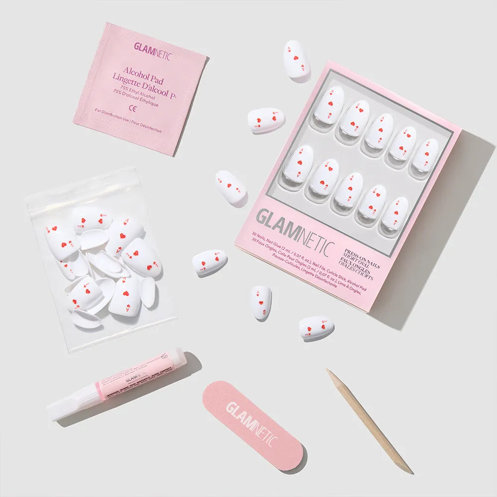 The Ace of Hearts by glamnetic is a white oval press-on nail set with small red hearts, including a pink file, wooden cuticle stick, nail glue, alcohol pad, and extra nails—all neatly displayed on a light gray surface. - Product image