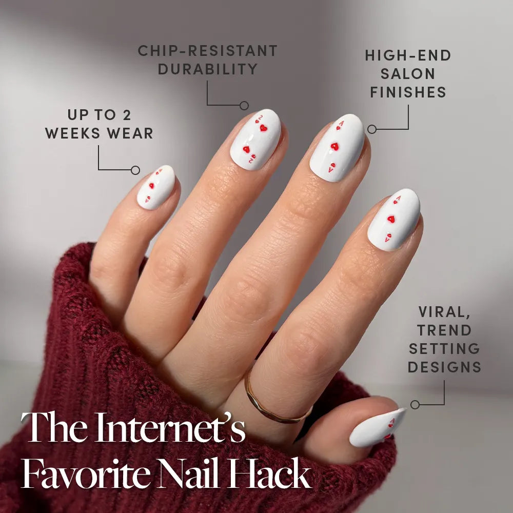 A hand wears glamnetic's Ace of Hearts oval press-on nails—white with small red hearts—and a burgundy sweater. Features include chip resistance, luxe finishes, up to 2 weeks’ wear, and trendsetting style. - Product image