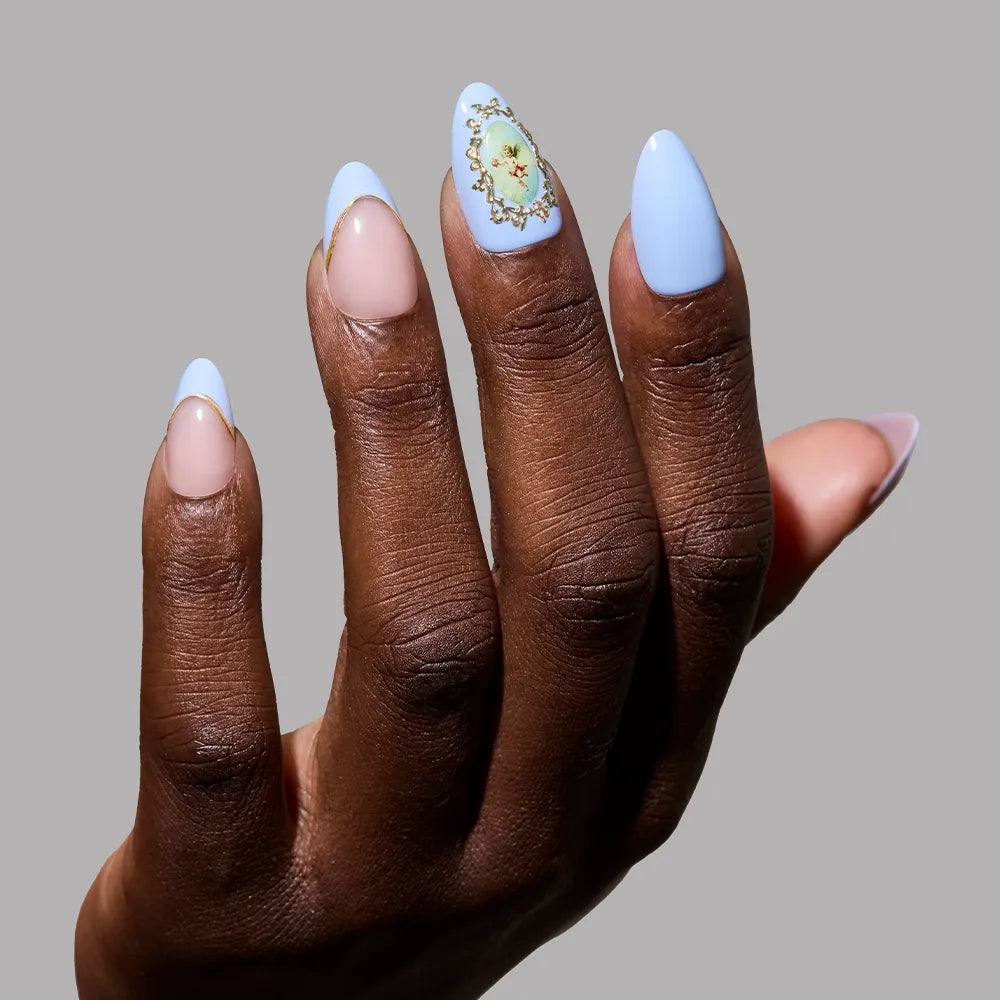 A hand with short almond nails features glamnetic’s Angel in Blue: three nails in light blue (one with a floral design), two in natural nude, and elegant 3D gold accents, all set against a plain gray background. - Product image