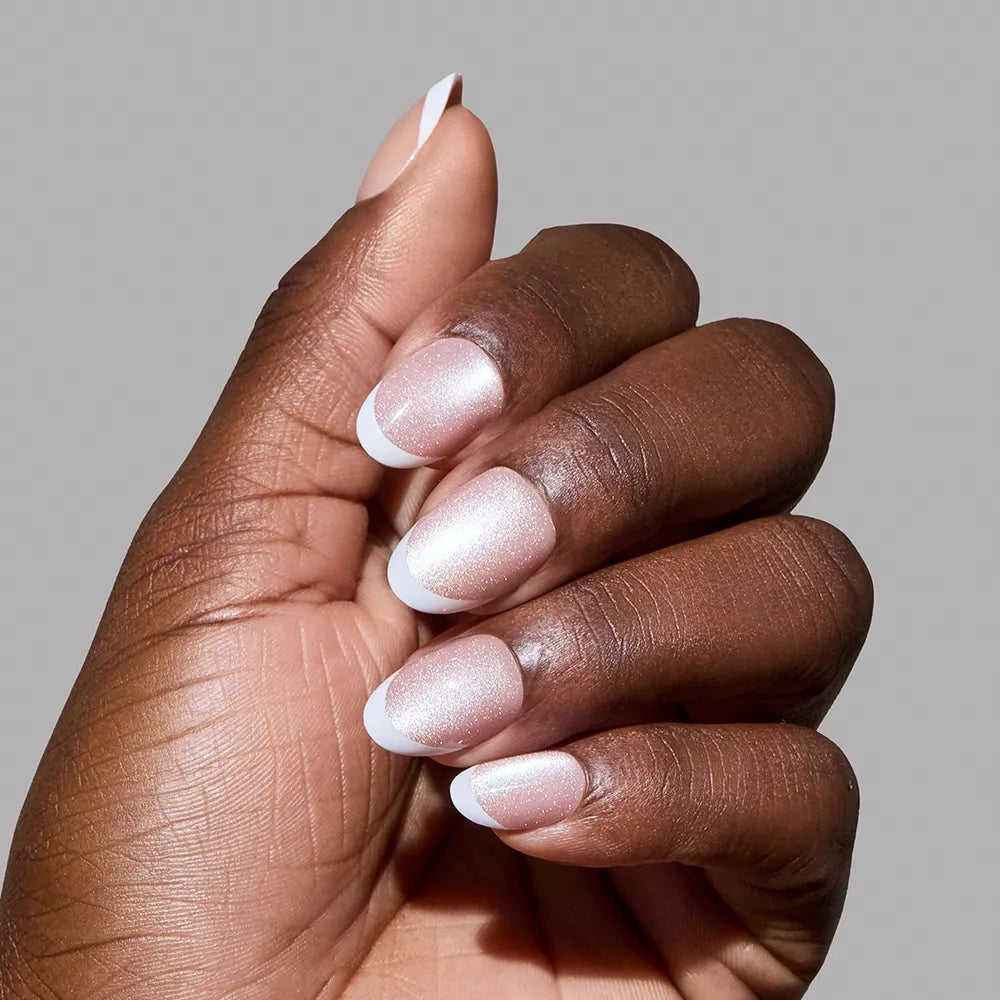 A hand with medium brown skin displays glamnetic's Angelic Touch: short oval nails in a nude velvet style with soft pink bases and crisp white French tips, set against a plain gray background. - Product image