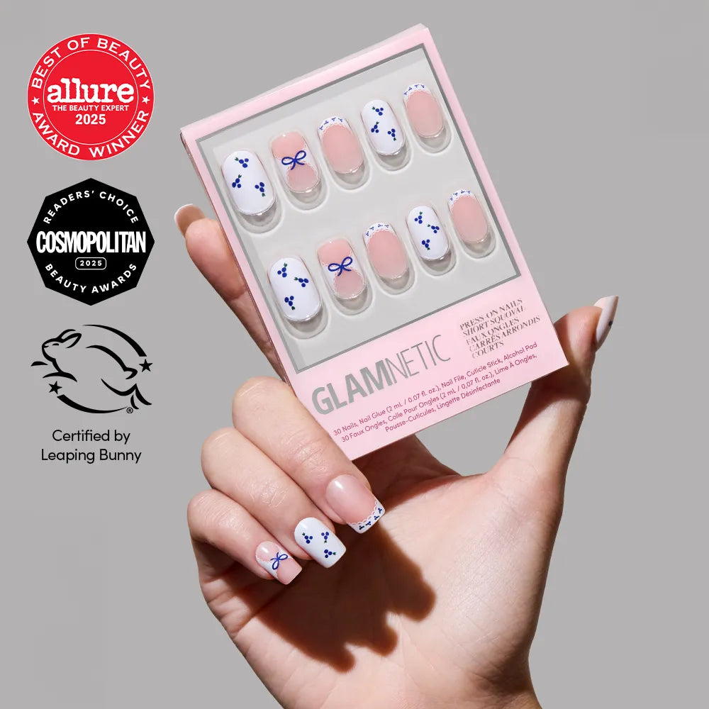 A hand with manicured squoval nails painted white and decorated with blueberry accents holds a box of glamnetic Blue A Kiss press-on nails. Beauty award logos are visible on the left.