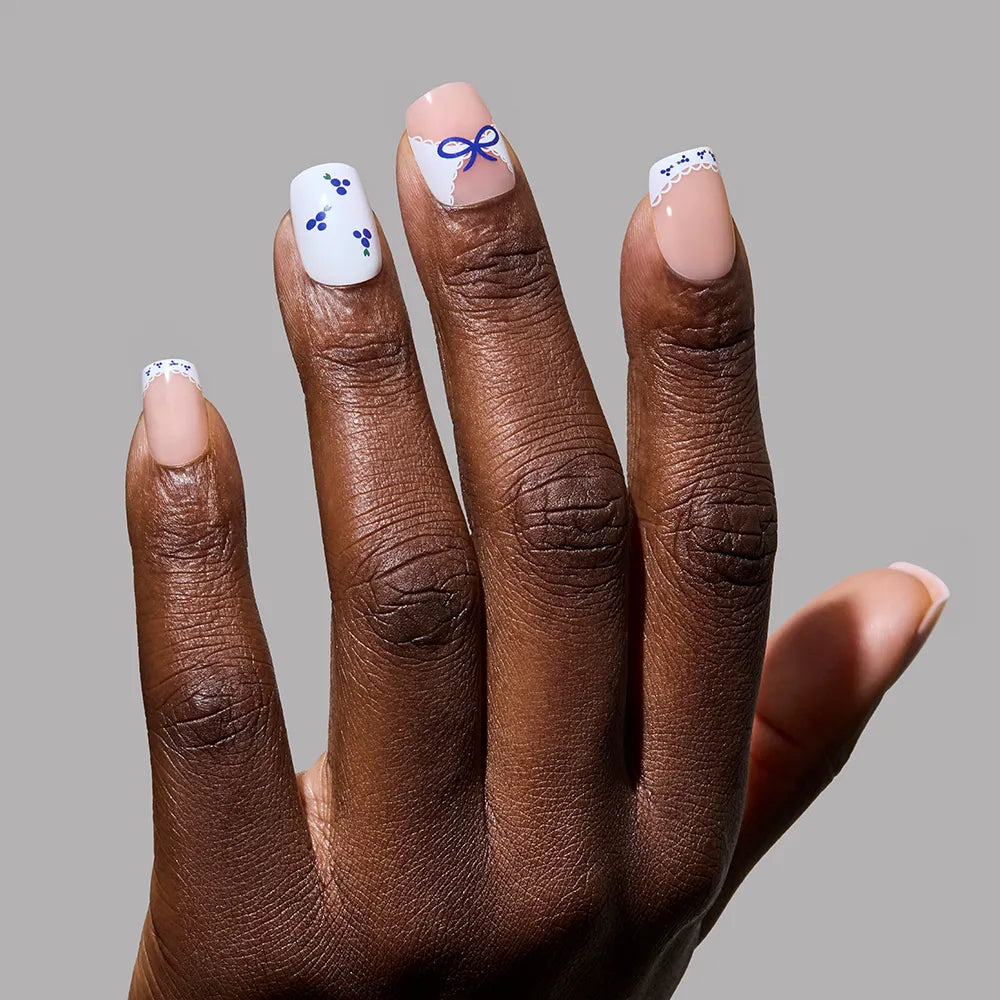 A manicured hand with glamnetic’s Blue A Kiss press on nails—squoval, nude base, and blueberry accents (dots, florals, bow on the middle finger)—set against a gray background. Perfect for anyone seeking unique nail charm. - Product image