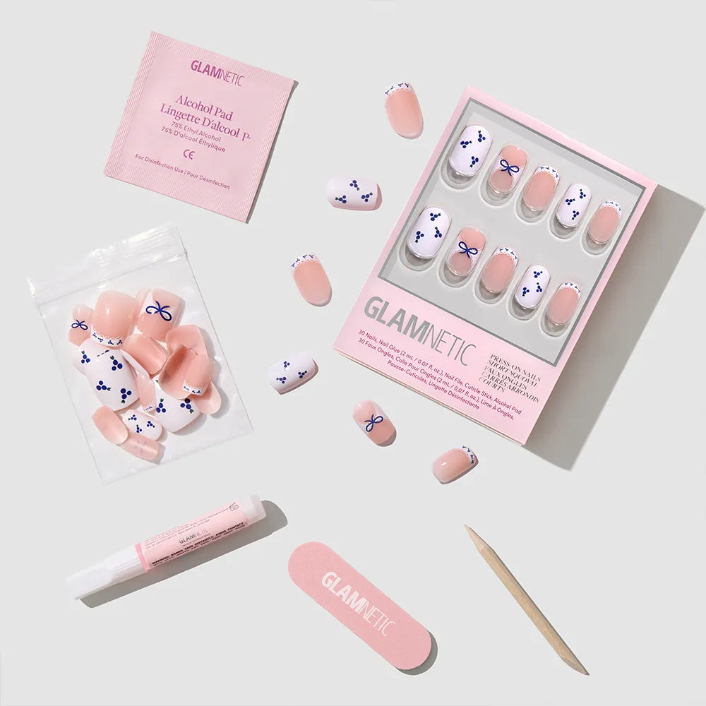 Blue A Kiss by glamnetic: squoval press-on nails with pink and blue bow designs and blueberry accents. Includes nail glue, alcohol pad, emery board, wooden cuticle stick, and resealable pouch, all displayed on a light surface. - Product image