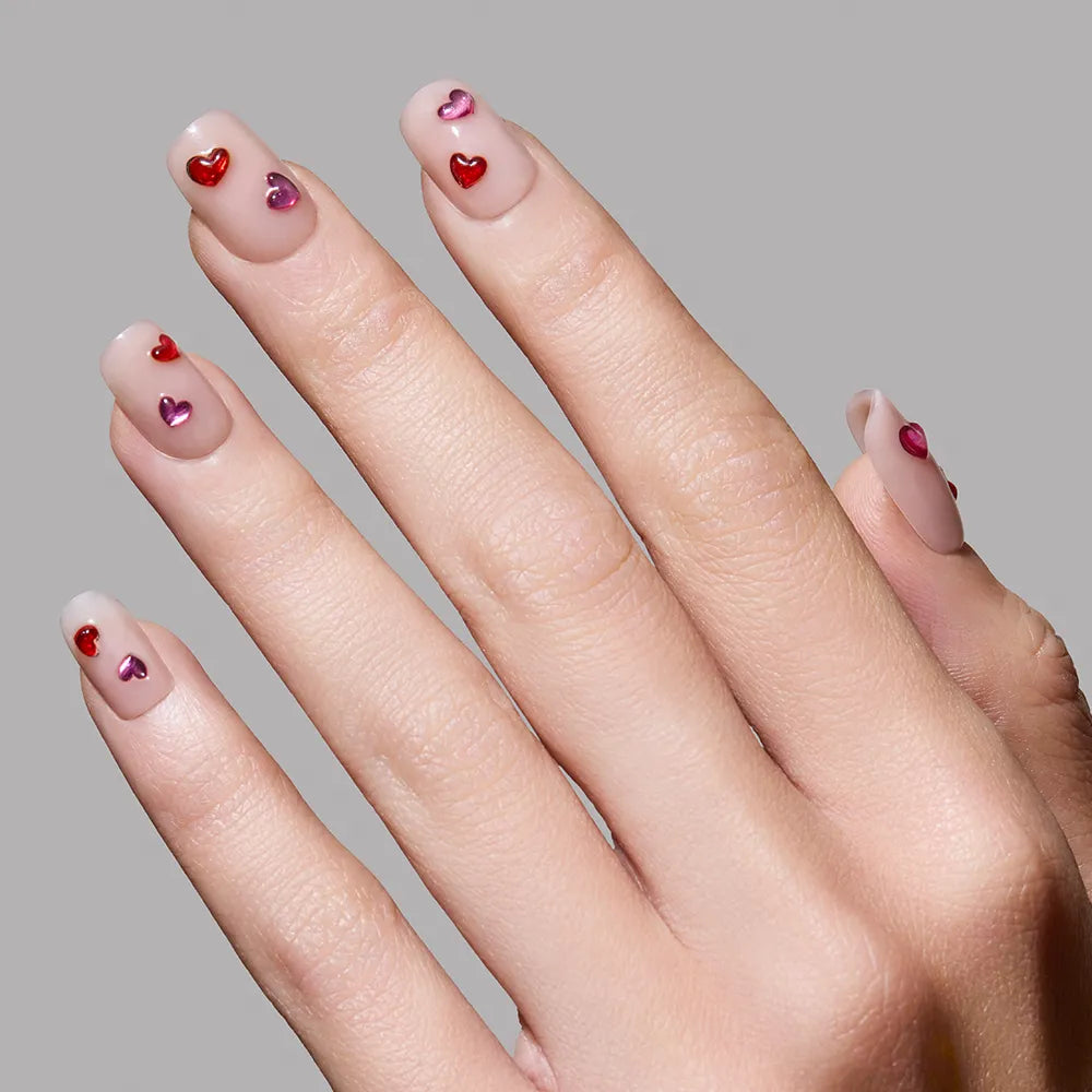 A hand showcases glamnetic’s Dear Lover nails: glossy nude, short squoval tips, each with shiny red and pink 3D gemstone hearts, set against a plain gray background. - Product image