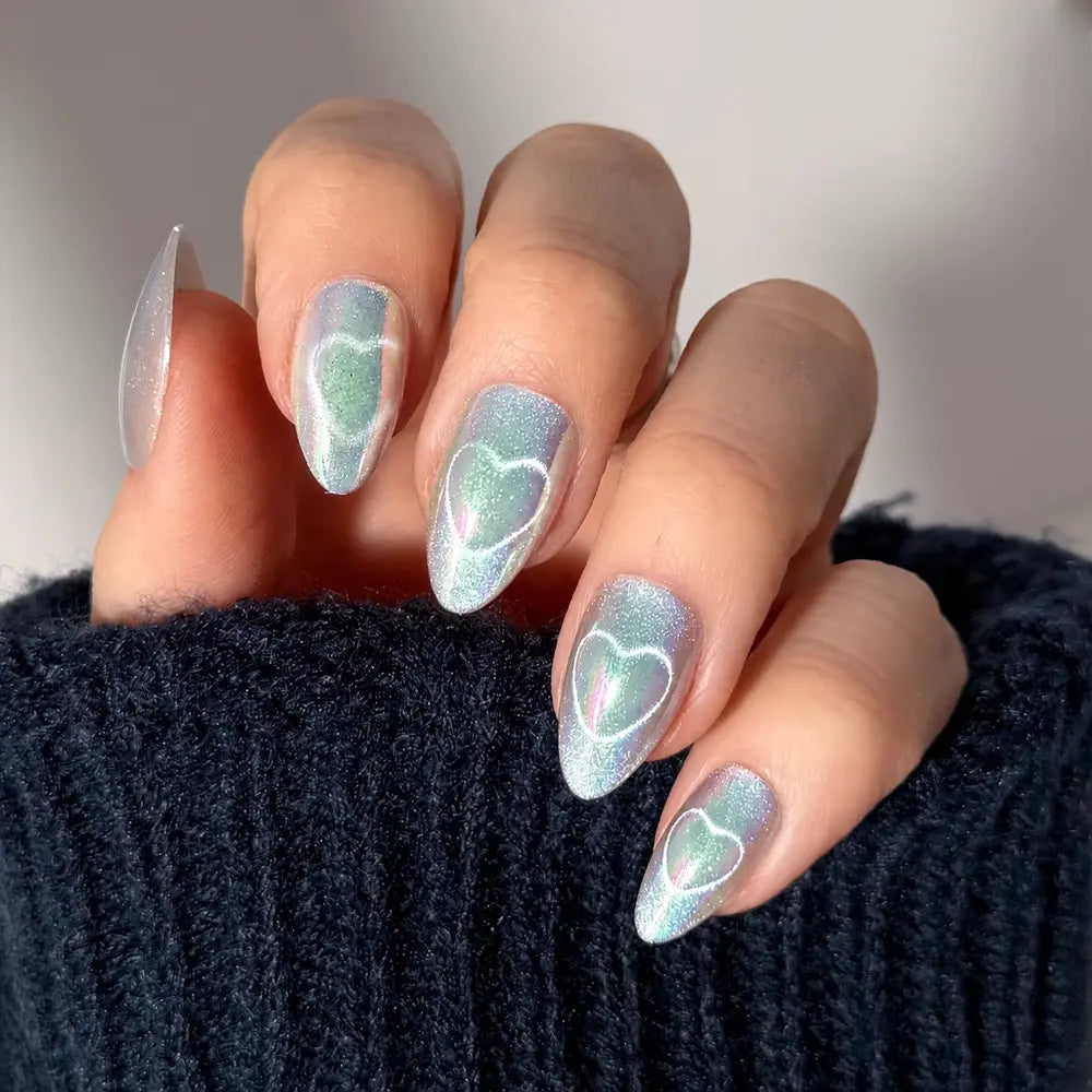 A hand with almond-shaped fairy-core nails painted in glamnetic’s Electric Lover, a shimmery iridescent light blue, each featuring a white heart outline. The person wears a dark, fuzzy sweater. - Product image