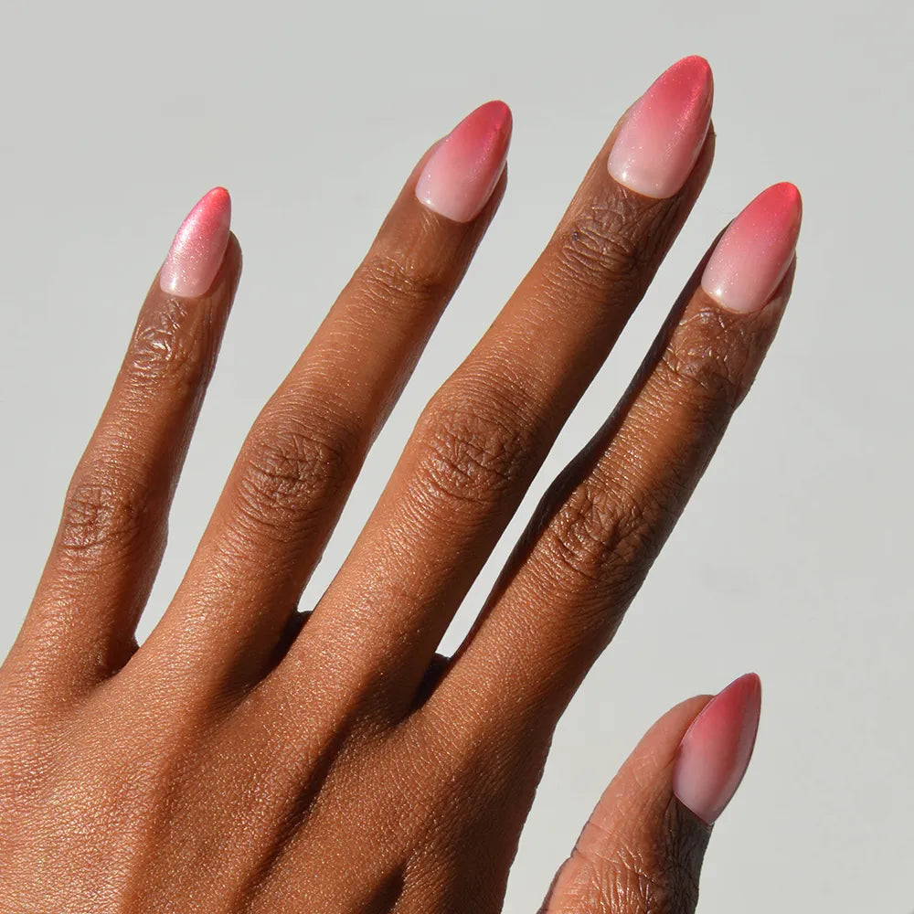 A hand with medium brown skin displays glamnetic Faded Feelings long almond press-on nails in a pink and white ombré against a plain light background.
