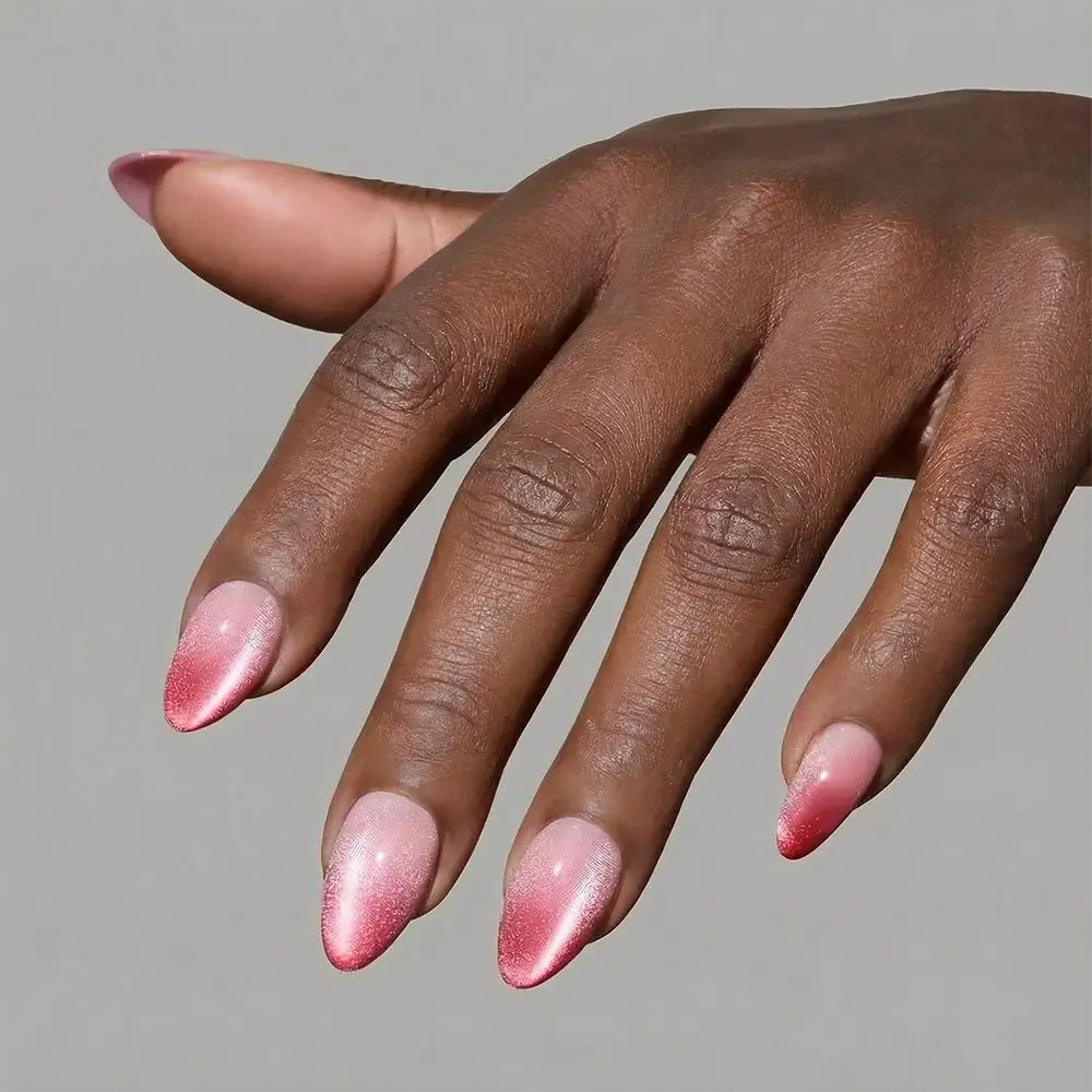 A hand with medium brown skin models glamnetic's Faded Feelings, long almond press-on nails featuring a pink shimmery ombré design, set against a plain gray background. - Product image