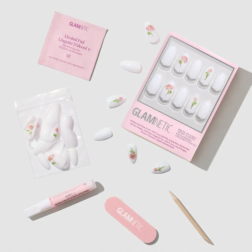 The glamnetic Floral Glow nail set features short almond nails with pink floral designs and 3D rose accents. It includes nail glue, a cuticle stick, a pink file, an alcohol pad, and extra nails displayed on a white surface. - Product image