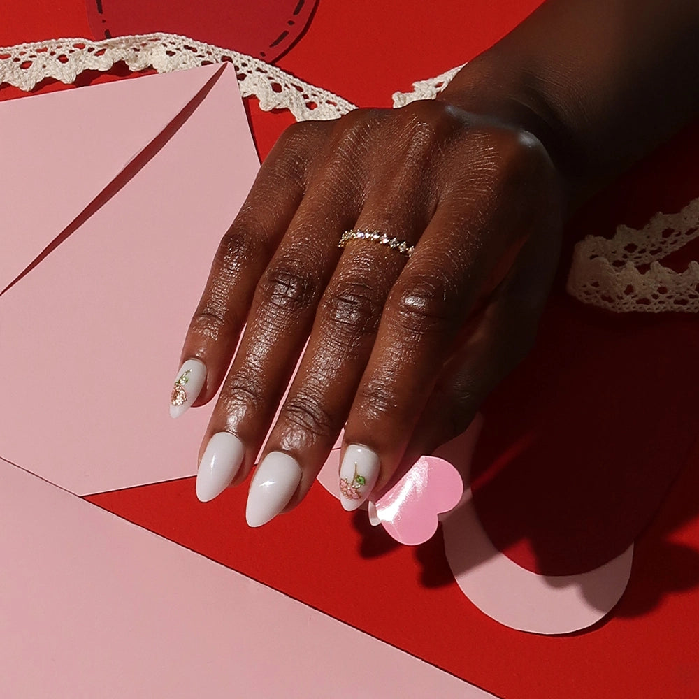 A hand with short almond nails from glamnetic’s Floral Glow set, featuring white tips and 3D rose accents, rests on pink paper with lace details; a delicate gold ring adorns the finger. - Product image