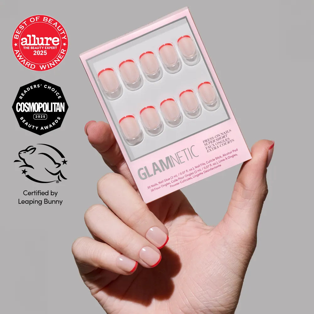 A hand holds a box of glamnetic Hotline Red press-on nails with red micro French tips and a glazed finish. Beauty award logos from Allure, Cosmopolitan, and Leaping Bunny certification are beside the product.