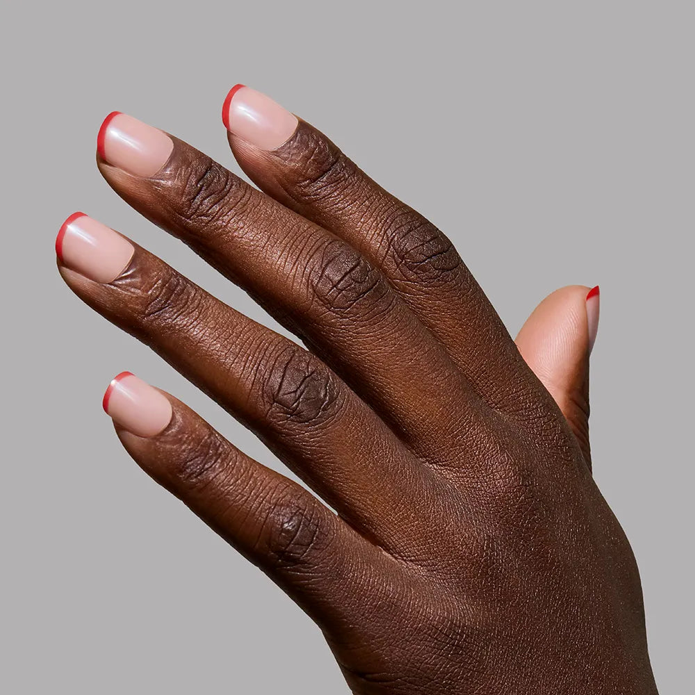 A hand with dark skin showcases neatly manicured nails with a nude base and red micro French tips using glamnetic's Hotline Red, set against a plain gray background. - Product image
