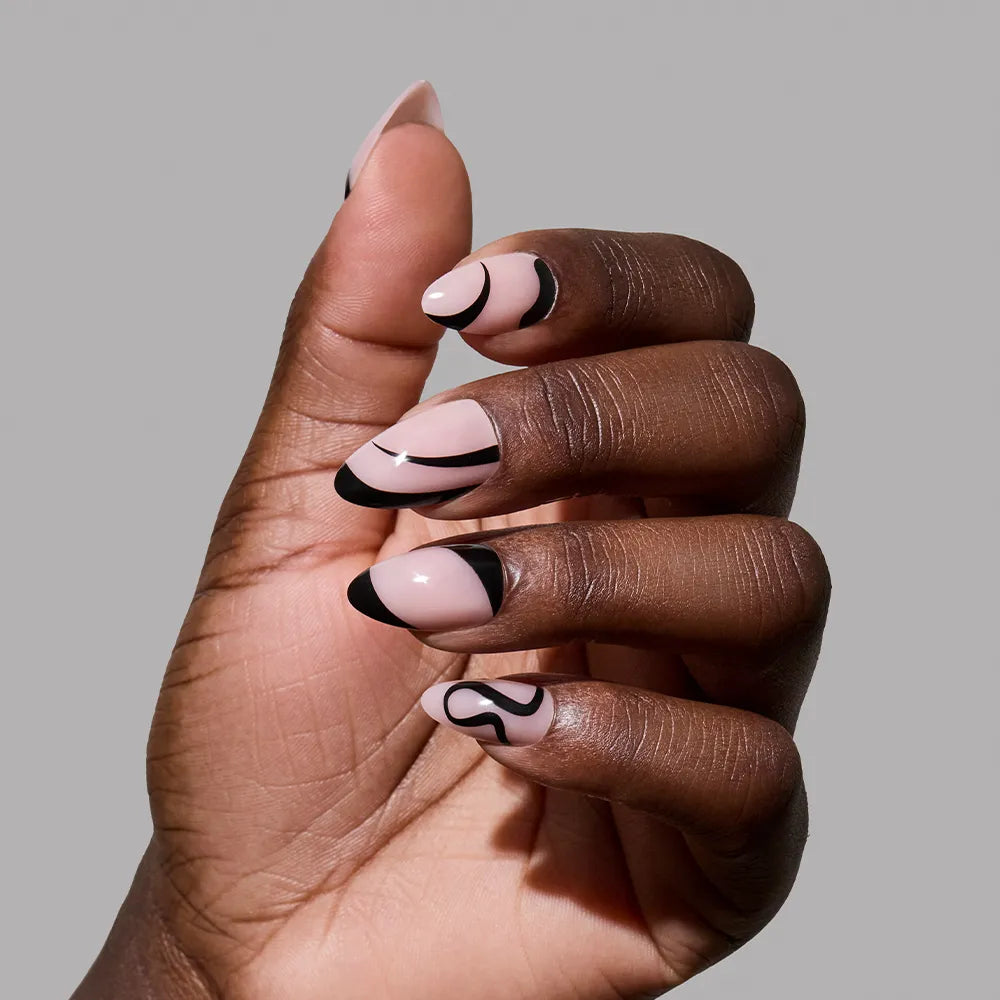 A hand with short almond nails wears glamnetic’s "Ink Wave" set—nude base nails detailed with black abstract lines and tips—highlighting a minimalist look against a plain grey background. - Product image