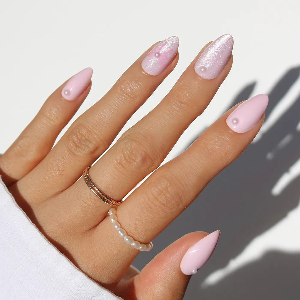 A hand wears glamnetic’s Love Knot long, almond-shaped pastel pink press-on nails with 3D pearl accents. Gold and pearl rings add to the soft-girl vibe, while a white sleeve contrasts the pristine background.