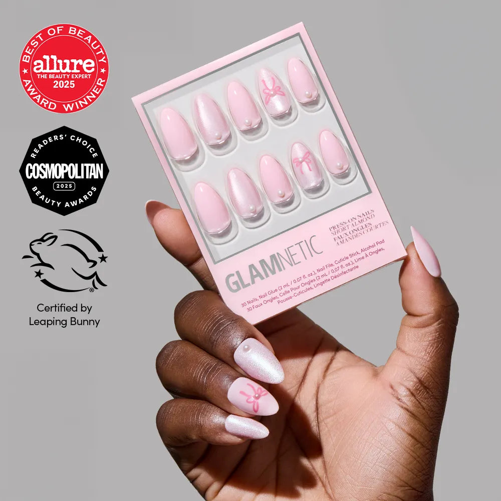 A hand with long, pale pink, glossy soft-girl nails holds a box of glamnetic Love Knot press-on nails. The packaging shows matching pink almond nails with a bow design, and various beauty award logos are featured next to the hand.