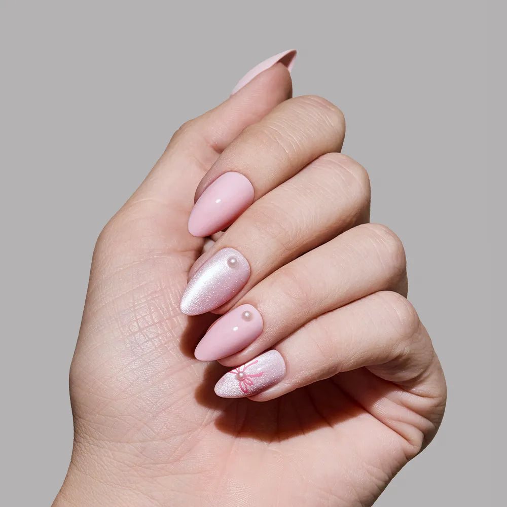 A hand wears glamnetic Love Knot almond press-on nails in soft pink with shimmer, 3D pearl accents, and a delicate red lace design on one nail, set against a plain gray background. - Product image