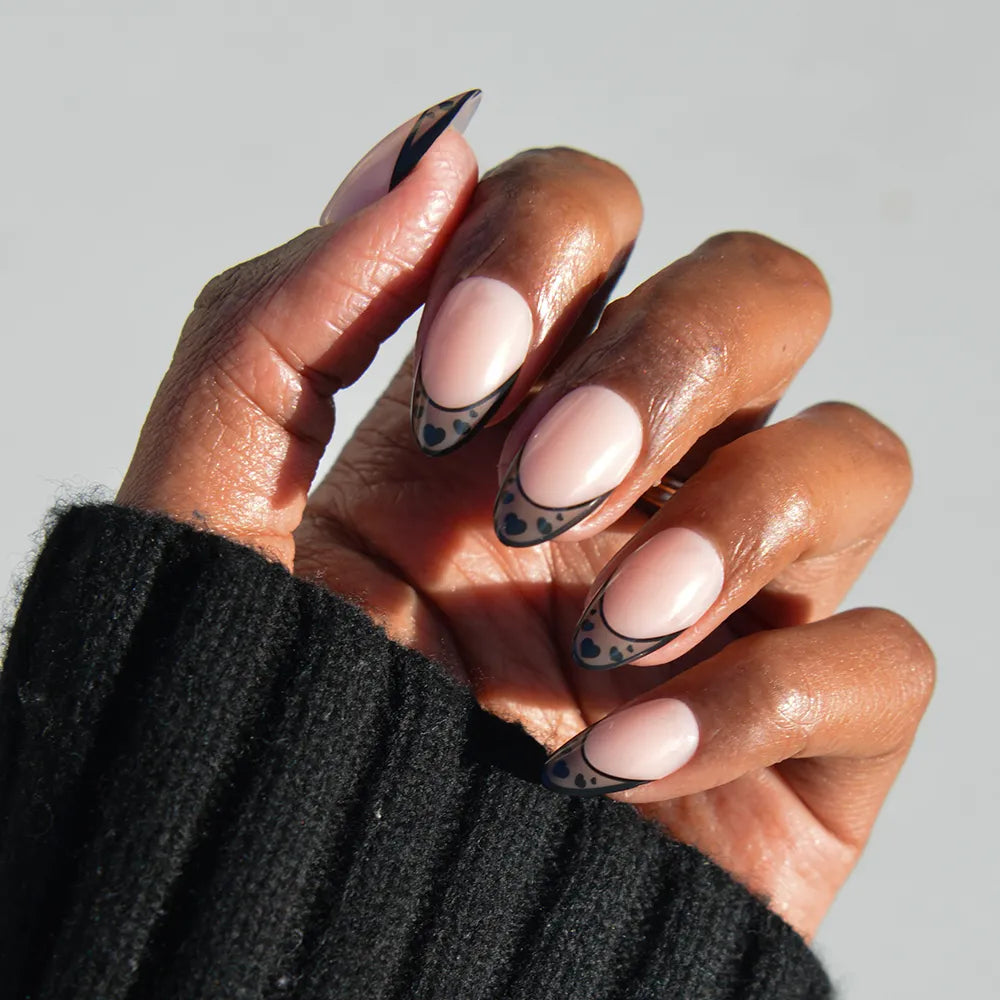 A hand with glamnetic's Love Noir short almond nails, featuring a nude base, black abstract French tips, and black dots, is shown against a light background. The wearer has on a black ribbed sweater. - Product image