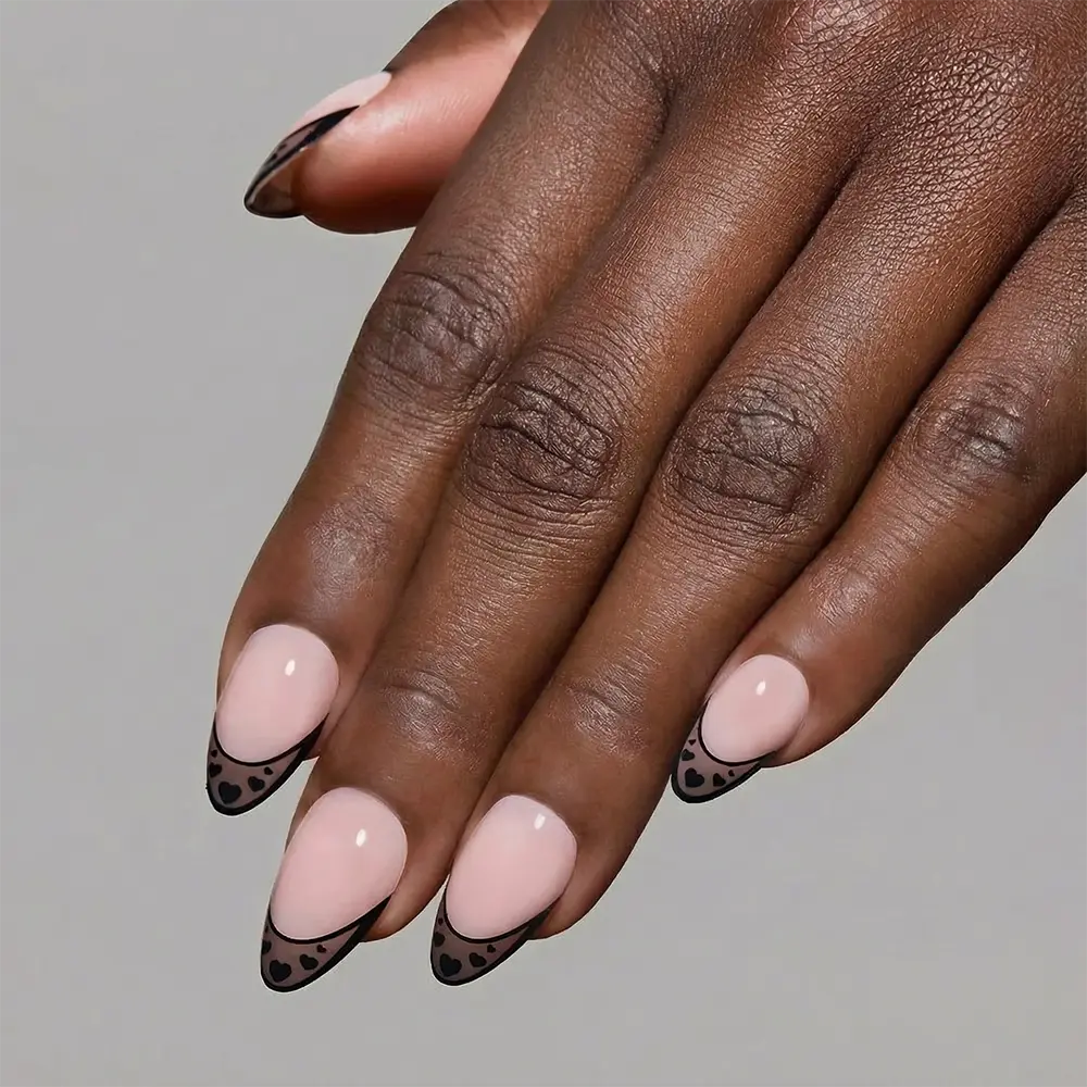 A hand with short almond nails, painted in glamnetic’s Love Noir—a nude shade with black French tips and tiny black hearts—displayed against a plain gray background. - Product image