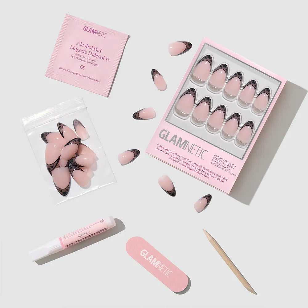 The glamnetic Love Noir set features pink and black press-on nails with a chic black heart design, a pink nail file, wooden cuticle stick, nail glue tube, alcohol pad, and extra nails—arranged neatly on a light gray surface. - Product image