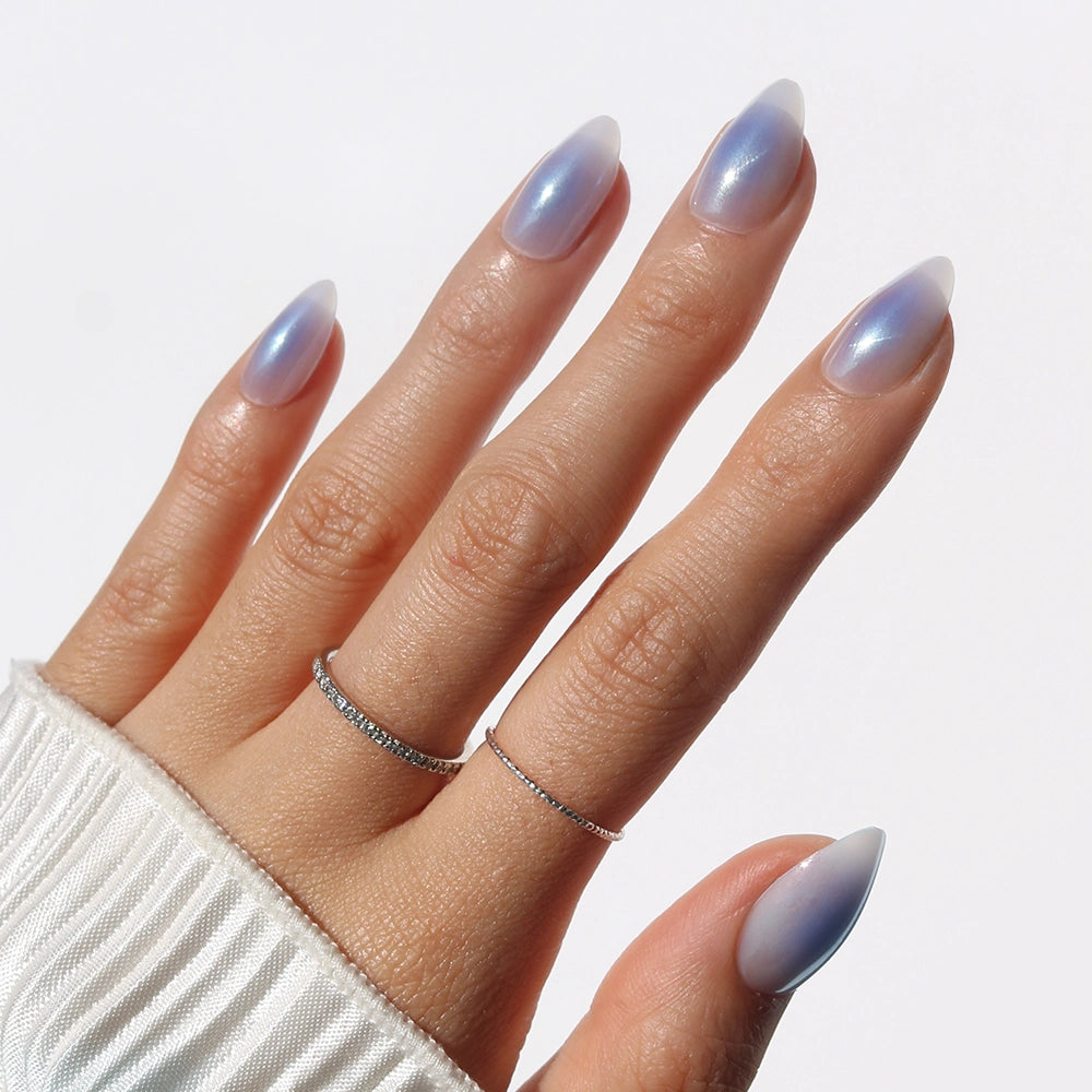 A hand with long, almond-shaped nails in glamnetic's Sky Berry, an iridescent light purple with a glazed finish. Two thin silver rings adorn the fingers, partially covered by a white ribbed sleeve against a plain white background.